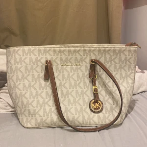 Beige väska från Michael Kors - Snygg beige väska från Michael Kors med MK-logga över hela väskan. Väskan har bruna handtag med gulddetaljer och ett MK-emblem. Insidan har flera fack och dragkedjor för smart organisering. Perfekt storlek för allt du behöver och stilren design. Dragkedjan är dock av men funkar att stänga 