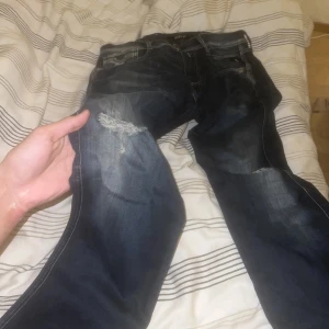 Mörkblå Replay jeans med slitningar - Säljer ett par mörkblå jeans från Replay med snygga slitningar på knäna. Jeansen har klassisk femficksdesign och tydliga kontrastsömmar. Perfekta för dig som gillar en lite ruffare streetstil.