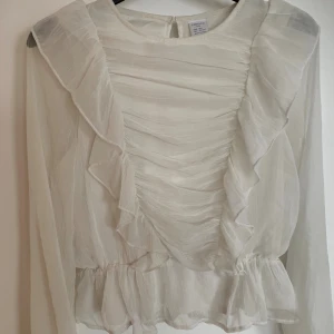 Vit volangblus från Lindex - Supersöt vit blus från Lindex med volangdetaljer längs axlarna och framtill. Blusen har långärmade, transparenta ärmar och en lätt rynkad struktur. Den är tillverkad i ett tunt, flowy material och har en liten volang vid midjan för extra feminin touch.