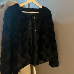 Svart pälsjacka från Vero Moda - Mysig svart pälsjacka från Vero Moda med långärmad design och rund halsringning. Jackan har knappar framtill och är tillverkad i mjukt fuskpälsmaterial som ger en lyxig känsla. Perfekt för att lyfta din outfit under kyliga dagar.