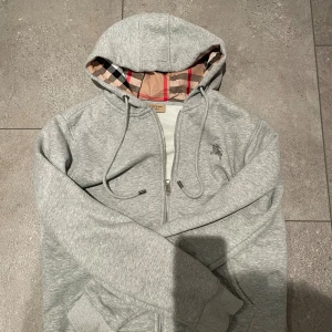 Grå Burberry hoodie med dragkedja - Snygg grå hoodie från Burberry i storlek M med klassiskt rutigt foder i huvan. Tröjan har dragkedja framtill, snörning vid huvan och Burberry-logga broderad på bröstet. Tillverkad i mjuk bomull, perfekt för chill dagar.