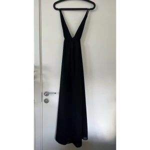 Svart långklänning från Favlook - Elegant svart långklänning från Favlook med djup v-ringning och smala axelband. Klänningen har en flowy passform och är tillverkad i ett lätt, tunt polyestertyg. Perfekt för dig som vill ha en stilren och tidlös look.