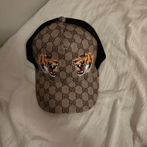 Gucci keps med tigertryck - Snygg keps från Gucci med klassiskt GG-mönster i brunt och två coola tigertryck framtill. Baksidan har svart mesh och justerbart band med gröna och röda detaljer. Perfekt för dig som vill sticka ut med lyxig streetstyle.
