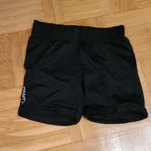 Svarta träningsshorts från Craft S - Svarta träningsshorts från Craft i storlek S. De är tillverkade i ett stretchigt syntetmaterial och har en elastisk midja samt en liten dragkedjeficka bak. Perfekta för dig som gillar att träna och vill ha bekväma shorts med sportig look.