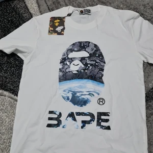Vit BAPE t-shirt med rymdtryck - Vit t-shirt från BAPE med ikoniskt ape-head tryck i rymdtema och jordmotiv. Stor BAPE-logga framtill. Klassisk passform och rund hals. Grym streetwear-vibe och perfekt för dig som gillar unika prints.