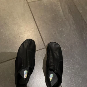 Svarta dansskor i skinn - Svarta dansskor från So Danca i mjukt skinn med elastiska partier på sidorna för smidig passform. Skorna har rund tå och platt sula, perfekta för dans eller gymnastik. Enkla att ta på och av tack vare stretchmaterialet.