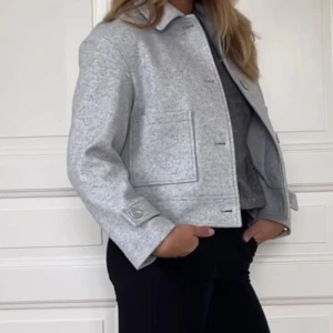 Grå kappa  - Stilren grå kappa  från 157 i ullmix med klassisk krage, stora fickor framtill och knäppning med stora knappar. Jackan har rak passform och är helfodrad för extra komfort. Perfekt för dig som gillar minimalistisk och clean stil. Trendig kappa! 