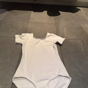 Vit kortärmad body med rund hals - En basic vit body med korta ärmar och rundad halsringning. Gjord i mjukt bomullsmaterial som sitter skönt mot kroppen. Perfekt att styla med jeans eller kjol för en clean look. Originellt balletbody.