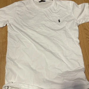  Polo Ralph Lauren - Vit klassisk t-shirt från Polo Ralph Lauren med broderad marinblå logga på bröstet. T-shirten har rund halsringning och korta ärmar. Tillverkad i mjuk bomull för en skön känsla och enkel stil.