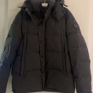 Svart dunjacka från Canada Goose M - Säljer en svart dunjacka från Canada Goose i storlek M. Jackan har en klassisk pufferstil med huva, dragkedja och knappar framtill samt två stora fickor med dragkedja. Perfekt för kalla vinterdagar och riktigt snygg streetstil.