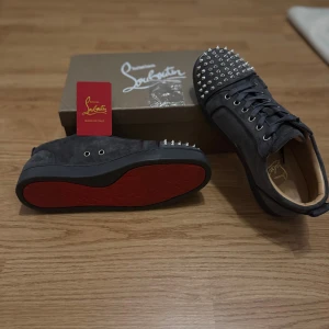 Christian Louboutin svarta sneakers med nitar - Svarta sneakers från Christian Louboutin i mocka med ikoniska röda sulor och silverfärgade nitar på tån. Skorna har snörning och rund tå, samt coola detaljer längs sidorna. Perfekt för dig som vill sticka ut med lyxig streetstyle.