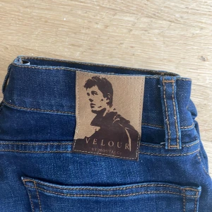 Mörkblå jeans från Velour by Nostalgia - Snygga mörkblå jeans från Velour by Nostalgia med klassisk femficksdesign och raka ben. Jeansen har kontrastsömmar och en stilren läderpatch bak i midjan. Perfekta för dig som gillar en enkel och tidlös look.