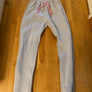 Ljusgrå mjukisbyxor från syna world storlek S - Säljer ett par ljusgrå mjukisbyxor från syna world med rosa snörning i midjan och broderad text 'Forgive Me' på benet. Baktill finns en stor rosa graffiti-logga. Byxorna har elastisk midja och är gjorda i mjukt bomullsmaterial.