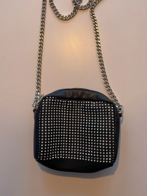 Svart axelväska med silverkedja - Snygg svart axelväska med glittriga paljetter på framsidan och en rejäl silverfärgad kedja som axelrem. Väskan har en rundad form och är tillverkad i skinnimitation. Perfekt för att lyfta din outfit med lite extra bling.