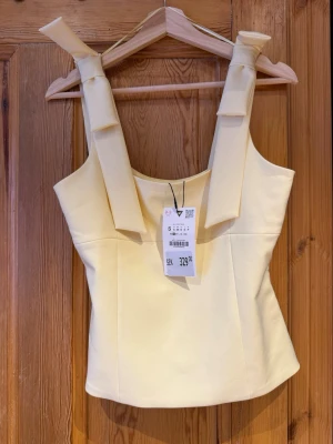 Ljusgul korsettopp med rosettband - Supersnygg ljusgul korsettopp från Zara med breda axelband som knyts till rosetter. Toppen har en figurnära passform och är tillverkad i ett slätt syntetmaterial som ger en stilren look. Perfekt för att lyfta din outfit med en feminin touch. Aldrig använt 
