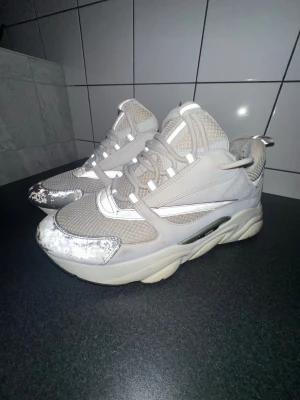 Dior B22 - Säljer ett par Dior B22 i vit och grå färg med mesh- och skinnpaneler. Skorna har snörning, platt sula och rund tå. Reflekterande detaljer på ovansidan och tjock yttersula ger en modern vibe. Perfekt för dig som gillar statement-sneakers.