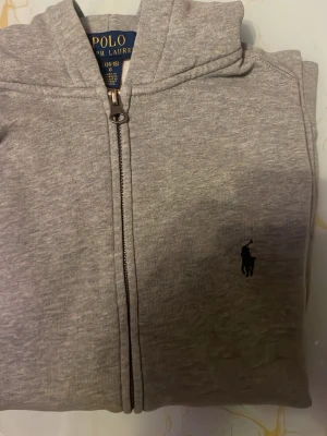Grå zip med huva från Polo Ralph Lauren - Grå zip med huva från Polo Ralph Lauren. Tröjan har dragkedja framtill, två fickor och en broderad logga på bröstet. Tillverkad i mjuk bomullsmix som känns skön mot huden. Perfekt för lager-på-lager och enkel att matcha med andra plagg.