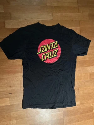 Svart Santa Cruz t-shirt med tryck - Svart t-shirt från Santa Cruz med stort, färgstarkt tryck på bröstet i rött och gult. Klassisk rund hals och korta ärmar. Materialet är mjuk bomull