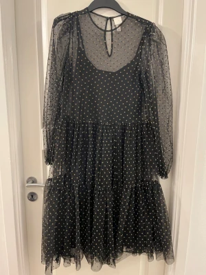 Svart prickig klänning från H&M - Säljer en svart kortklänning från H&M med transparenta långärmar och små beige prickar över hela klänningen. Klänningen har volangdetaljer och är gjord i ett lätt syntetmaterial som ger en luftig känsla. Perfekt för dig som gillar trendiga och lekfulla plagg.