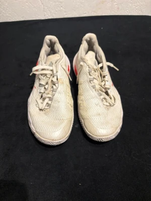 Adidas vita sneakers med röda detaljer - Säljer ett par vita Adidas sneakers med röda och silvriga detaljer på sidorna och sulan. Skorna har snörning och är tillverkade i syntetmaterial med mesh-överdel för bra ventilation. Yttersulan har ett mönstrat grepp och klassiska Adidas-ränder på sidan.