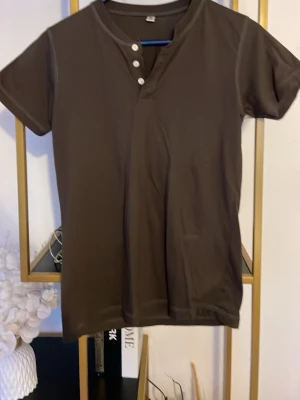 Brun t-shirt med knappar i halsen - Säljer en brun t-shirt med korta ärmar och snygg knappdetalj vid halsen. T-shirten har en enkel och stilren design utan mönster, perfekt för dig som gillar basic plagg med en twist. Materialet känns mjukt och bekvämt mot huden.