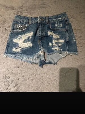 Blå jeansshorts med nitar från Bik Bok - Säljer ett par blå jeansshorts från Bik Bok i storlek XS. De har råa, fransiga kanter och slitna detaljer framtill. Ena fickan är dekorerad med silvriga nitar för en cool touch. Klassisk femficksmodell med knapp och dragkedja.