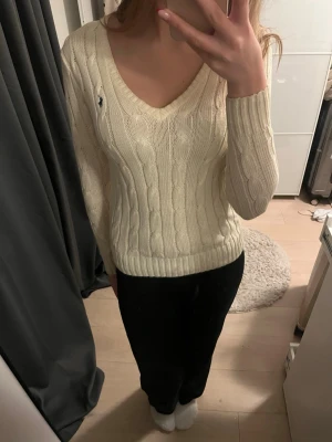 Vit kabelstickad tröja från Ralph Lauren - Säljer en klassisk vit kabelstickad tröja från Ralph Lauren med v-ringning och lång ärm. Tröjan har den ikoniska lilla broderade loggan på bröstet och är gjord i mjuk bomull. 