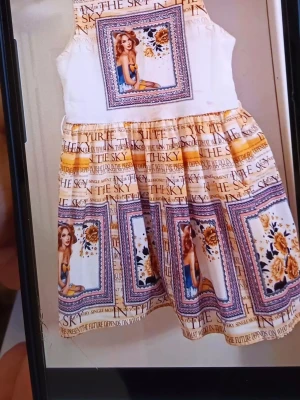 Färgglad kortklänning med tryck - Unik kortklänning med färgglada tryck av texter, blommor och illustrationer. Klänningen har en vit överdel och mönstrad kjol i nyanser av gult, blått och beige. Ärmlös modell med rund halsringning och markerad midja. Perfekt för dig som gillar statement-plagg.