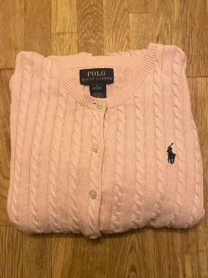 Rosa kofta från Polo Ralph Lauren - Snygg ljusrosa kofta från Polo Ralph Lauren. Den passar storlekarna 146, 152 och 158 och säker mer och mindre! Den har klassiskt kabelstickat mönster och knappar framtill. Tröjan har rund hals, långa ärmar och den ikoniska broderade loggan på bröstet. Perfekt för dig som gillar stilrena och tidlösa plagg❤️