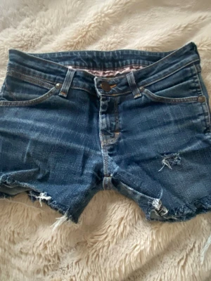 Mörklblå jeans shorts från Wrangler! - Säljer ett par blå jeansshorts från Wrangler som tidigare var jeans med klassisk femficksdesign och rå, fransig kant nedtill. Shortsen har låg midja och slitna detaljer framtill samt tydliga W-sömmar på bakfickorna. Perfekta för sommarens chill dagar. Ställ gärna frågor 🥰