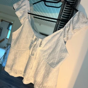 Vit topp med volangdetaljer - Säljer en superfin vit offshoulder topp med volangdetaljer. Toppen har en söt knytning framtill och är perfekt för sommardagar. Materialet är lätt och luftigt, vilket gör den idealisk för varma väder. Passar perfekt till både jeans och kjol!