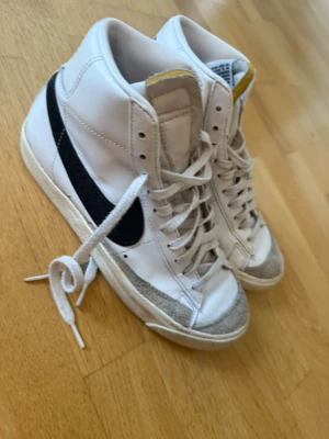Nike Blazer Mid vita sneakers - Nike Blazer Mid sneakers i vitt med svart swoosh på sidan. Höga med snörning och rund tå, ovandel i skinn och detaljer i mocka vid tån. Klassisk retrostil med tydlig Nike-logga på hälen och platt sula. Perfekt för dig som gillar streetwear och sportig vibe.