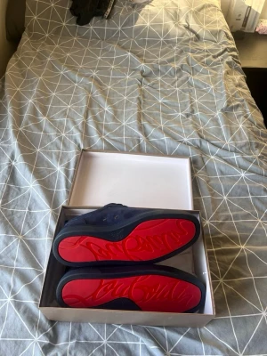 Mörkblå sneakers från Louboutin - Säljer ett par mörkblå sneakers från Louboutin med snygg röd sula och broderad logga på hälen. Skorna har snörning och är tillverkade i mocka med platt sula. Kommer i originalkartong och har en riktigt lyxig vibe.