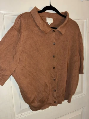 Brun kortärmad kofta från H&M - Kortärmad brun kofta från H&M i mjukt stickat material. Klassisk krage och knäppning med bruna knappar framtill. Perfekt till lager på lager och enkel att styla med olika outfits. Passar dig som gillar en chill och enkel look.