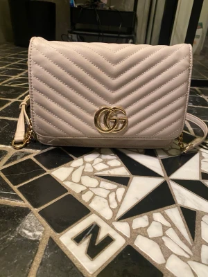 Beige axelväska  i skinn - Säljer en beige axelväska  i mjukt skinn med quiltad design och guldfärgad GG-logga framtill. Väskan har justerbar axelrem med kedjedetaljer och flera fack inuti, perfekt för att hålla ordning på dina grejer. Stilren och klassisk modell som lyfter varje outfit.