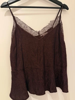 Vinröd spetsdetaljerad linnetopp H&M - Säljer en vinröd linnetopp från H&M i storlek 38. Toppen har tunna axelband, spetsdetaljer vid halsringningen och ett diskret paisleymönster i tyget. Materialet är mjukt och lätt, perfekt för varma dagar eller lager på lager. Snygg och feminin look!