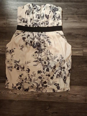 Blommig axelbandslös kortklänning - Snygg axelbandslös kortklänning med svart midjeband och blommigt mönster i svart och grått på en ljus, nästan vit bakgrund. Klänningen har en figurnära passform och är fodrad med mjukt tyg. Stängs med dragkedja i ryggen och har fina veckdetaljer vid midjan.Har även fickor på båda sidorna 