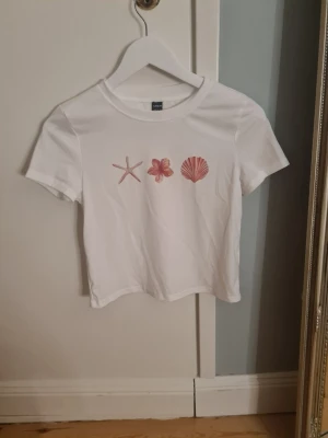 Vit t-shirt med strandmotiv från SHEIN - Vit t-shirt från SHEIN i bomull med korta ärmar och tryck framtill av en sjöstjärna, en blomma och en snäcka i rosa toner. Enkel och fräsch design som passar perfekt till sommaren. Rund halsringning och normal passform.