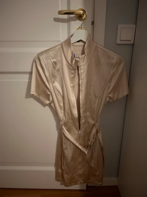 Beige satin klänning med bälte - Snygg beige kortklänning från Selected i glansigt satinmaterial. Klänningen har korta ärmar, hög krage, dragkedja framtill och ett brett bälte med metallspänne i midjan. 