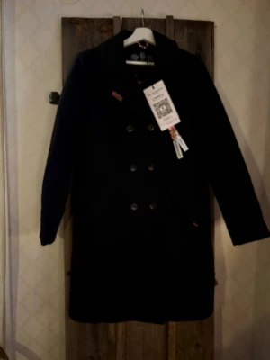 Klassisk kappa/ rock från Navahoo  - Säljer en klassisk och varm svart kappa/ rock från Navahoo, i färgen svart. Längre modell med dubbelknäppning framtill, reverskrage, istoppsfickor i sidan samt innerficka/ fickor. Perfekt för kalla dagar!