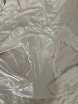 Vit långärmad topp med spetskant - Säljer en vit långärmad topp från Brandy Melville med söt spetskant längs den vida halsringningen. Toppen är i mjuk bomull och har en slim passform som sitter snyggt på kroppen. Perfekt att styla med jeans eller kjol för en enkel och fräsch look.