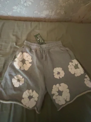 Denim tears shorts - Grå shorts från denim tears med elastisk midja och snörning. De har stora vita blommönster tryckta över hela tyget och är tillverkade i mjuk bomull. Passformen är loose och längden går till knäna, perfekt för chill dagar.