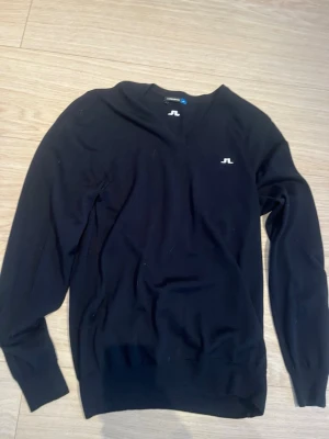 Svart stickad tröja från J.Lindeberg - Säljer en svart stickad tröja från J.Lindeberg i storlek S. Tröjan har v-ringning, långa ärmar och vita J.Lindeberg-loggor på bröstet, ärmslutet och nacken. Enkel och stilren design som passar perfekt till lager på lager under höst och vinter. 100% merino ull