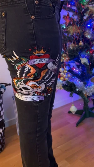Svarta lowaist ed hardy jeans - Säljer ett par svarta jeans från Ed Hardy med färgstarkt broderi på benet som visar en döskalle, hjärta och texten 'New York City'. Bakfickan har röd Ed Hardy-logga och läderpatch med logotyp. Skinny fit men utsvängda. De är low waist💗De finns två små hål. Priset kan diskuteras