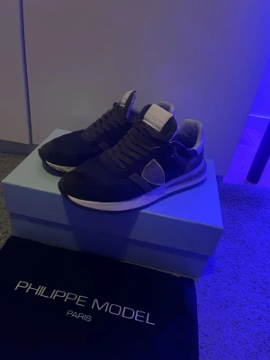 Philippe Model sneakers i mocka - Säljer ett par Philippe Model sneakers i mörkgrön och svart mocka med vita detaljer och bruna snören. Klassisk låg modell med rund tå och vit sula. Skorna har den ikoniska sköld-loggan på sidan och en bekväm innersula. Perfekta för dig som gillar stilrena och exklusiva sneakers. Skorna är sparsamt använda och nypris ligger på 4-5 tusen. Storlek 40 