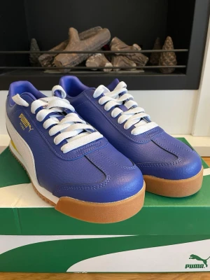 Blå Puma Roma sneakers - Säljer ett par Puma Roma sneakers i blått skinn med vit sula och vita snören. Skorna har detaljer i gult och brunt på sulan, samt klassisk Puma-logga i guld på sidan. Snygg retrostil med rund tå och platt sula.