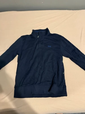 Marinblå half zip tröja från BOSS - Snygg marinblå långärmad tröja från BOSS med half zip och hög krage. Tröjan har diskret BOSS-logga på bröstet och ribbade muddar vid ärmslut och nederkant. Perfekt för dig som gillar stilrena och klassiska plagg med sportig touch.