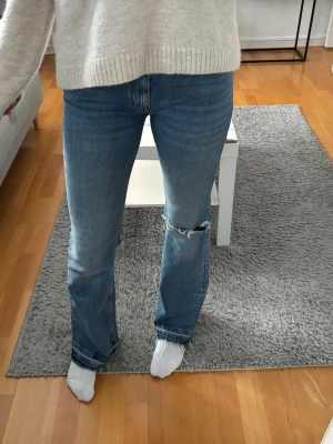 Blåa jeans med hål - Säljer ett par lager157 jeans i klassisk blå tvätt med bootcut passform. Jeansen har ett stort hål med slitningar på ena knät och råa, fransiga benslut för en cool vibe. Materialet är robust denim och midjan är normal. Perfekt för dig som gillar en avslappnad och trendig stil. Byxorna är nersprättade längst ner för att de skulle bli längre.