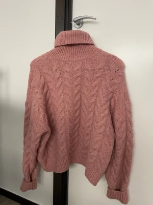 Rosa polotröja med flätstickning - Mysig rosa polotröja med grov flätstickning och ribbade muddar. Tröjan har lång ärm och hög krage som ger extra värme. Perfekt för kyliga dagar och har en loose passform som känns avslappnad och trendig.