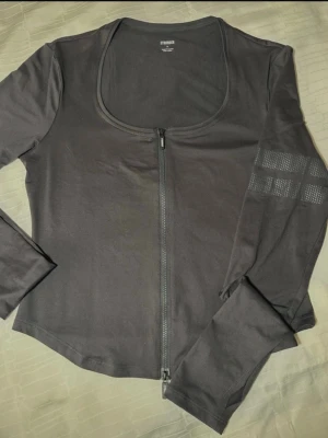 Grå  zip-tröja från Stronger - Grå  långärmad tröja från Stronger med rundad hals och hel dragkedja framtill. Tröjan har diskreta detaljer på ena ärmen och är tillverkad i mjukt, stretchigt syntetmaterial som passar perfekt till träning. Snygg och enkel design med logga i nacken. Användes en gång bara. 